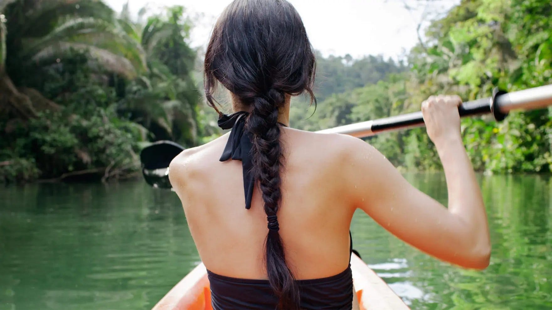 Mujer con cabello largo y negro en una trenza navegando en kayak por un río de espaldas a la cámara